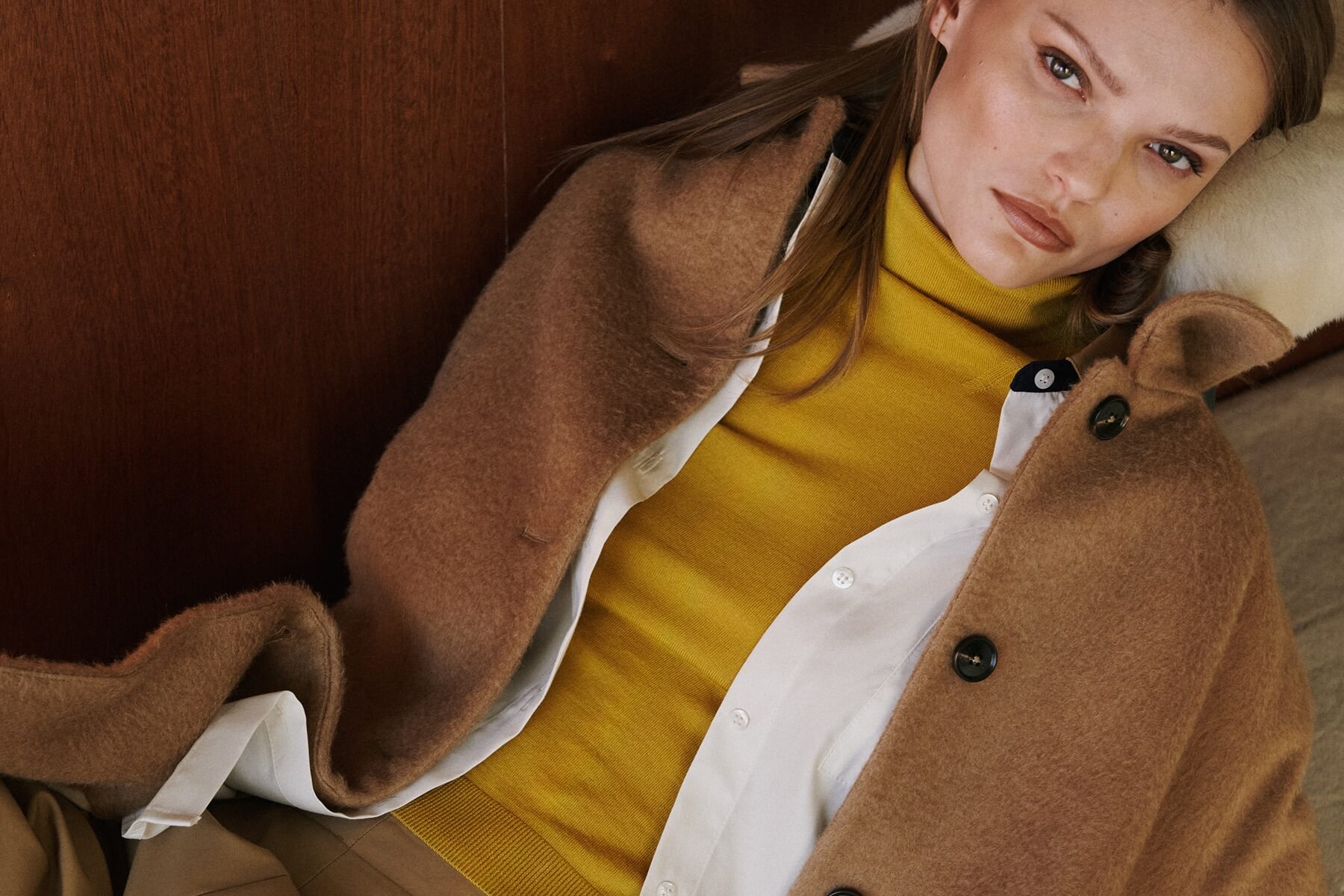 Close up of woman wearing layered camel colored coat over yellow turtleneck and white button down.