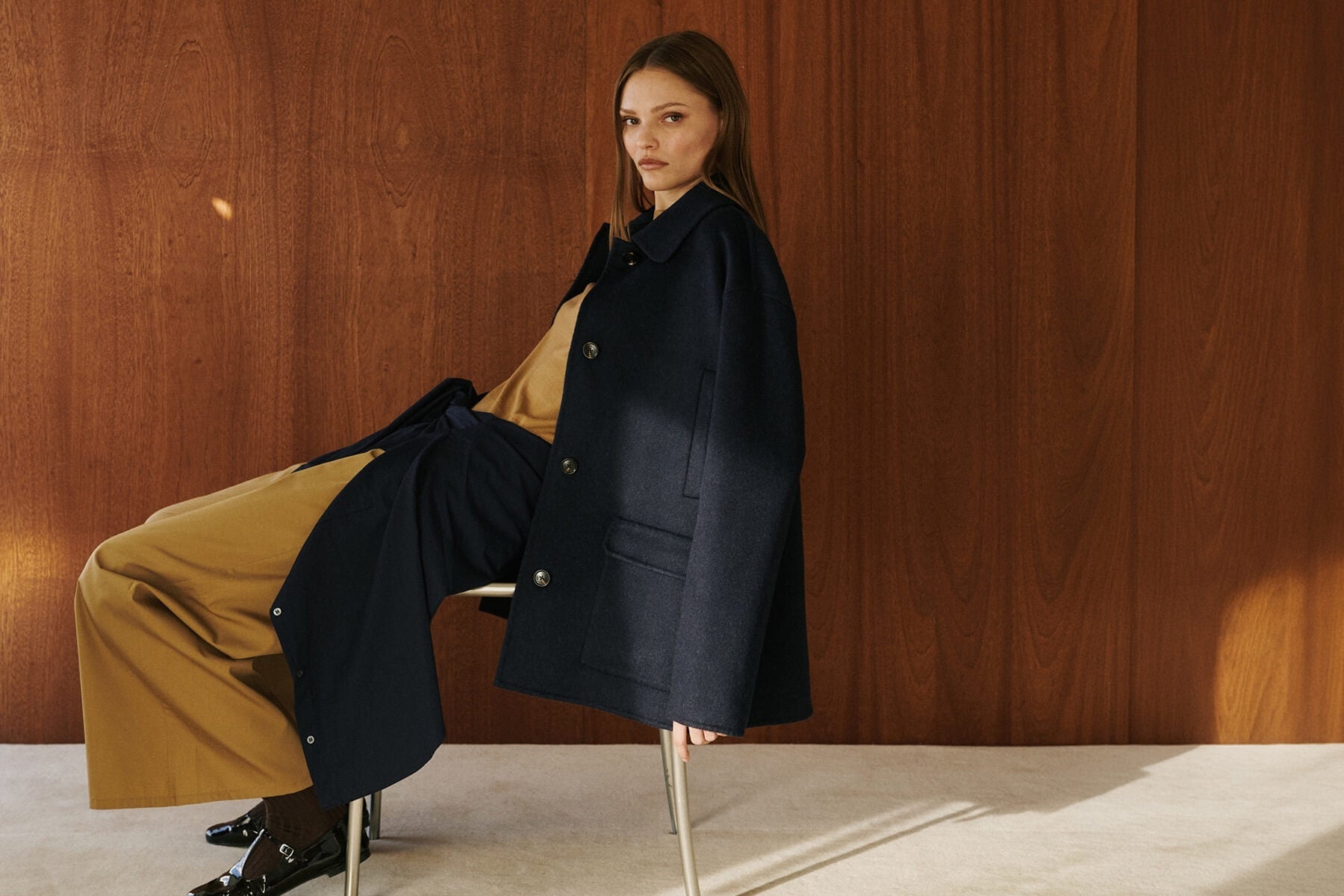 Woman sitting in camel colored pants with black coat and sweater draped over.