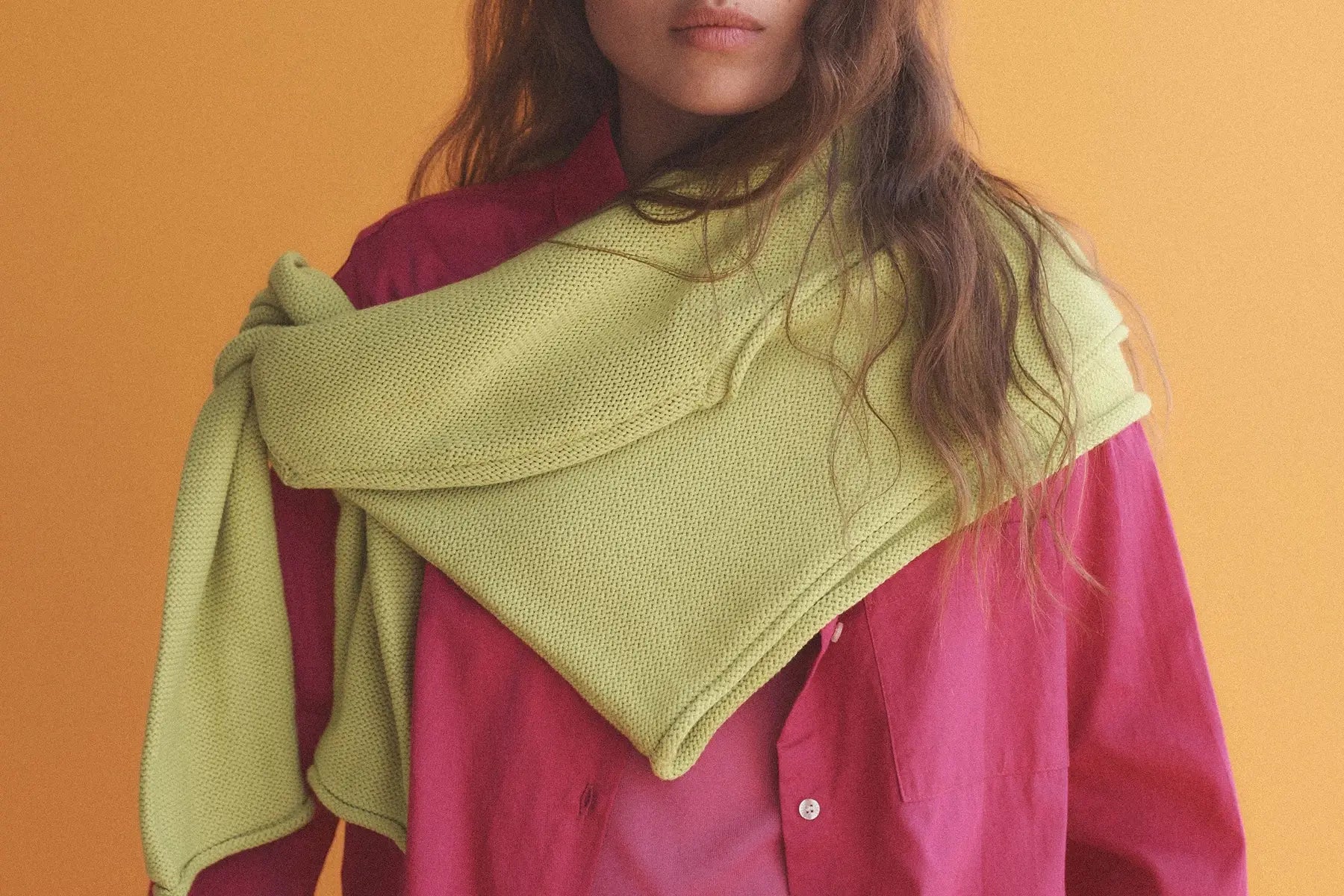 Close up of woman wearing chartreuse sweater draped over pink button down, with orange background.