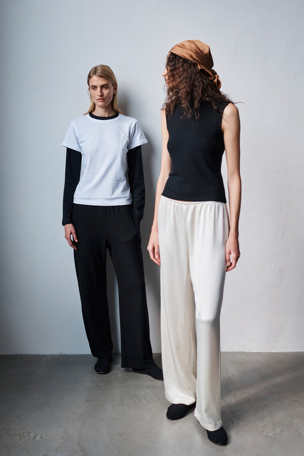 Two women standing against wall in barb satin pants, with layered tshirt and sleeveless crew, looking off into distance.