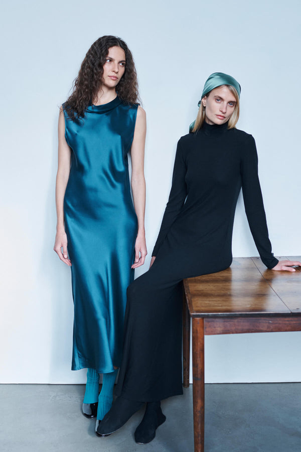 Two women in maxi dresses, one leaning against the table in black, the other in pacific satin