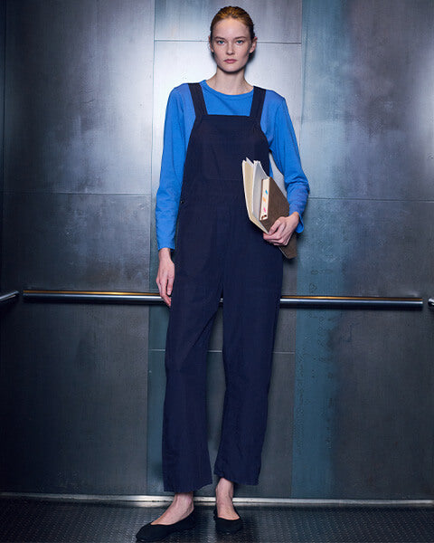 Woman walking forward carrying books, in front of glass window with city view, wearing crop trousers with black flats and white and black long sleeve tshirt.