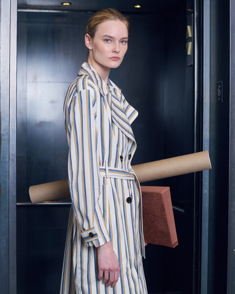 Woman standing in stripe trench coat holding a rolled cardboard and binder, in front of elevator.