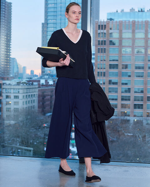 Woman walking forward carrying books, in front of glass window with city view, wearing crop trousers with black flats and white and black long sleeve tshirt.