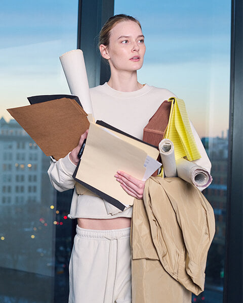 Woman standing in front of city view window in chalk colored Jamie sweat suit, holding binders, folders and notepads and a coat.
