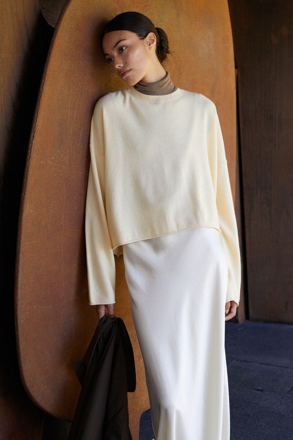 Woman leaning on wall, in yellow crewneck sweater and white silky skirt, holding jacket