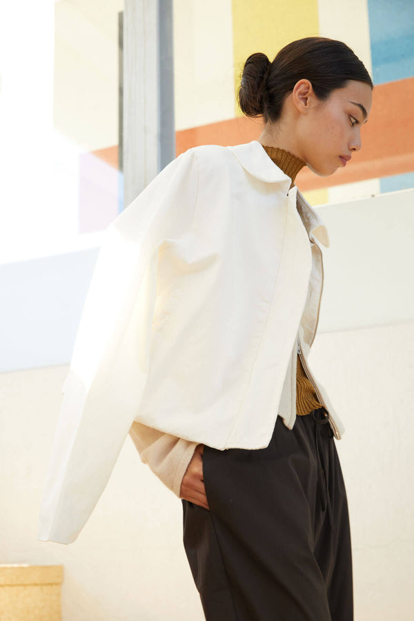 Woman in white zip up jacket and brown trousers, hands in pockets