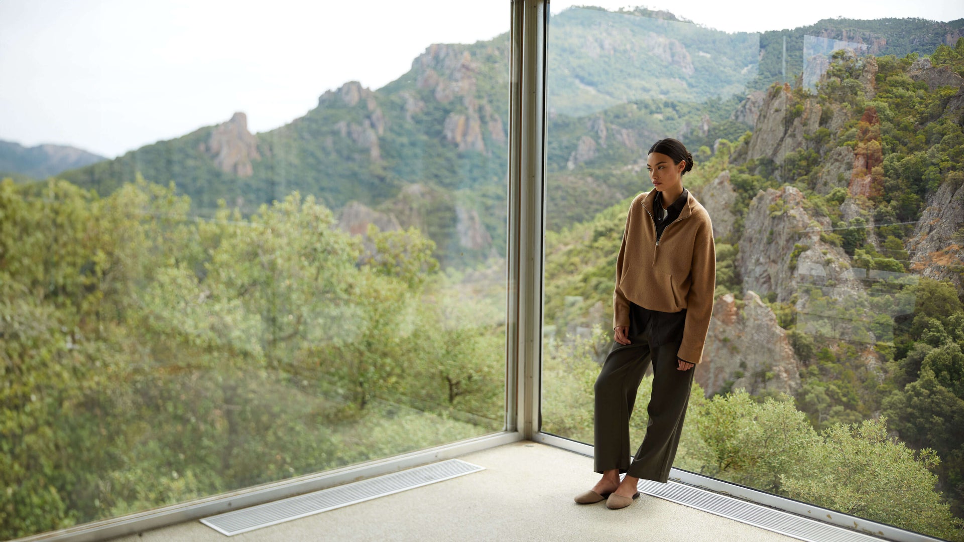 Woman standing by window with scenic view in Jess anorak camel and Chloe olive pants