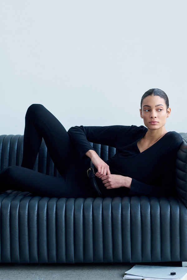 Woman laying on couch, in black long sleeve v neck and leggings, looking to the side.
