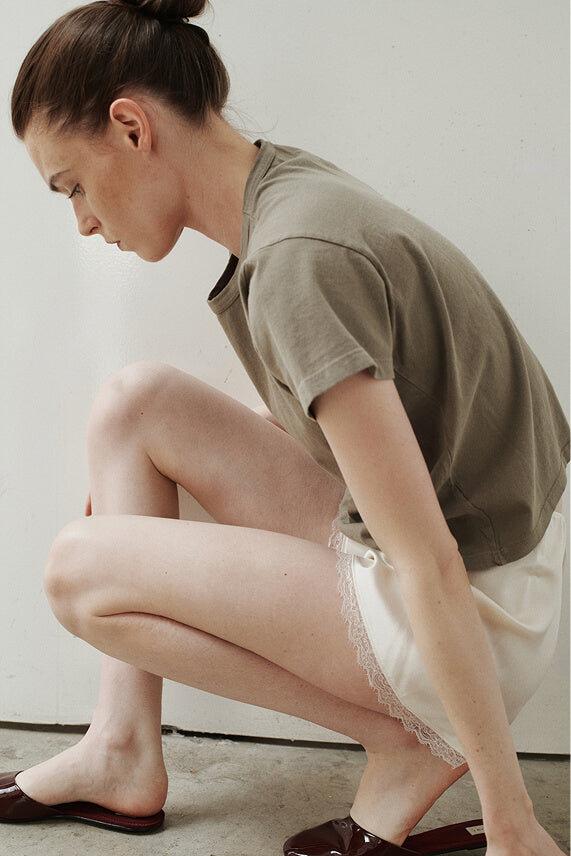 Woman squatting down in lace mini skirt and cypress tshirt in front of blank wall.