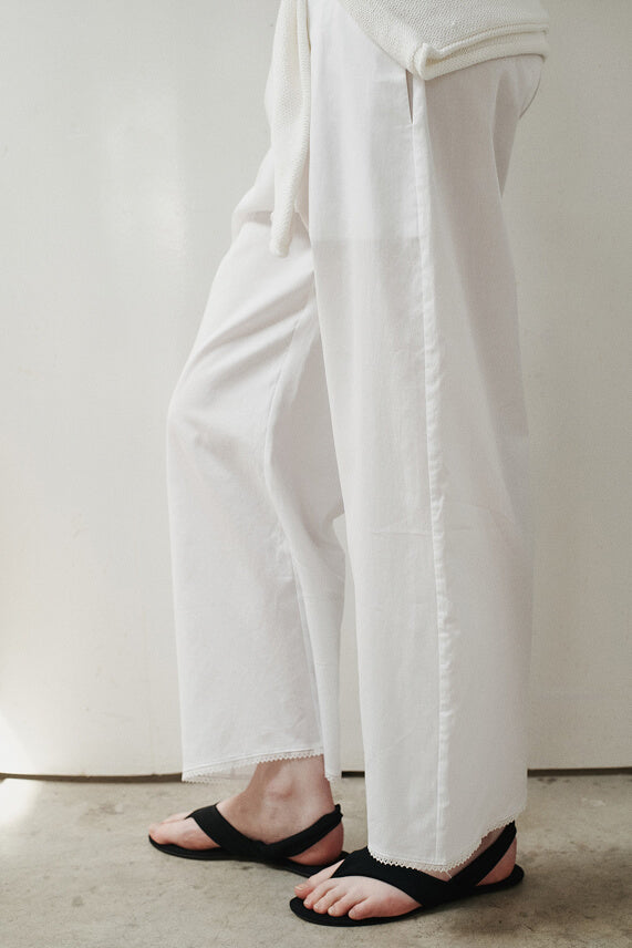 Close up of white cotton lace trim pants, with black sandals, standing in front of blank wall.