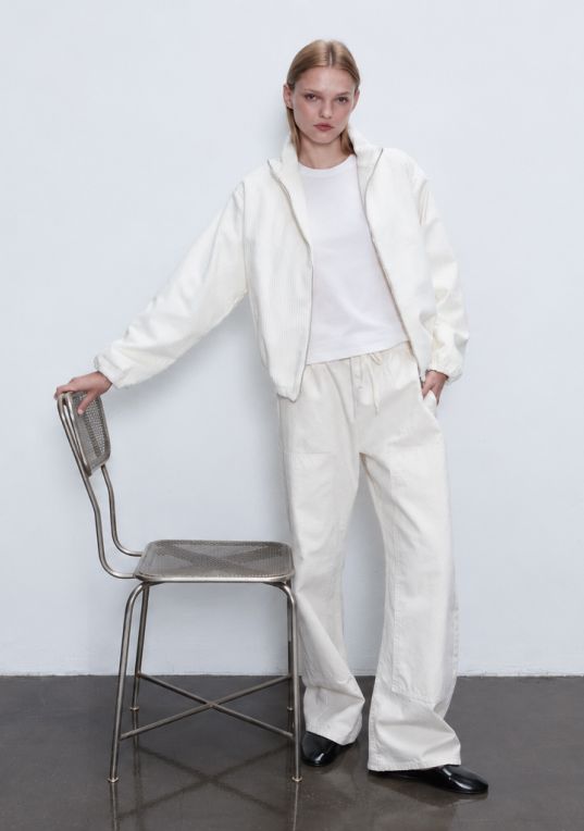 Model is standing in white tee and sammi corduroy white jacket and pants holding metal chair.