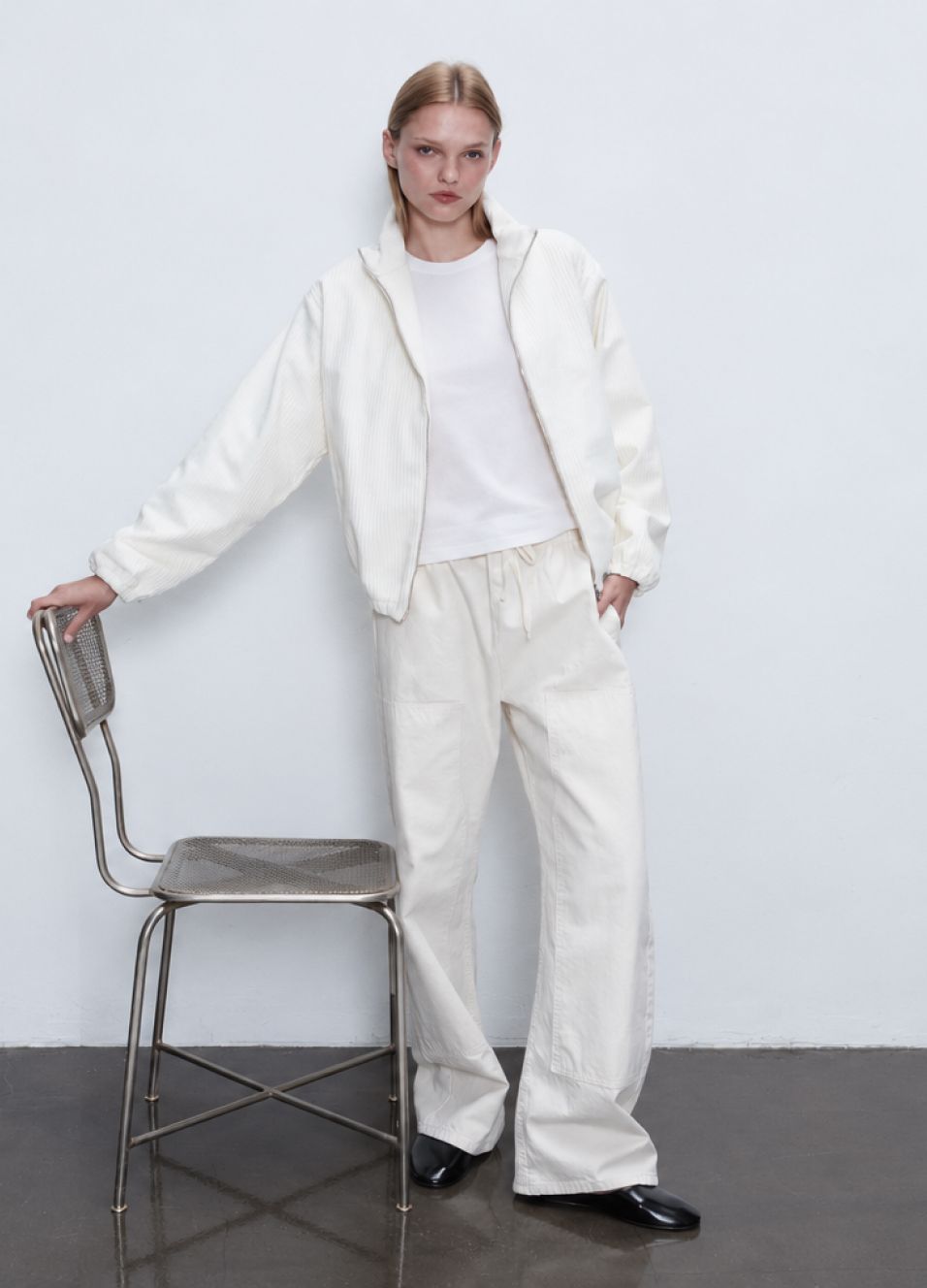 Model is standing in white tee and sammi corduroy white jacket and pants holding metal chair.