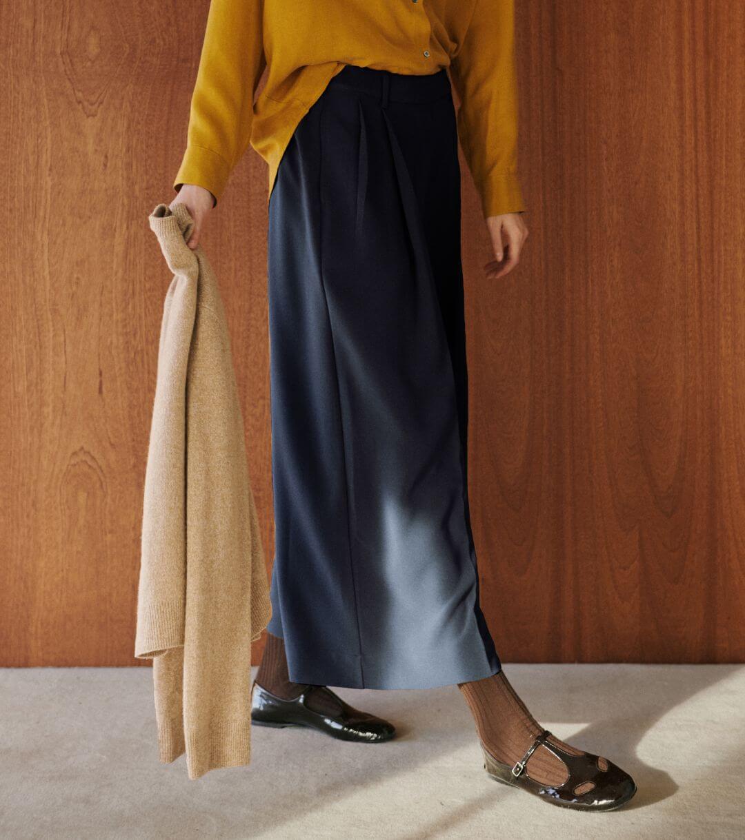 Close up of pleated pants on model with brown mary janes and socks, holding camel sweater.