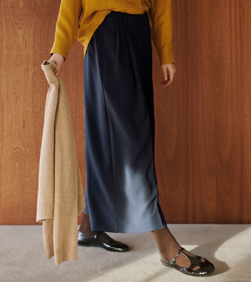 Close up of pleated pants on model with brown mary janes and socks, holding camel sweater.