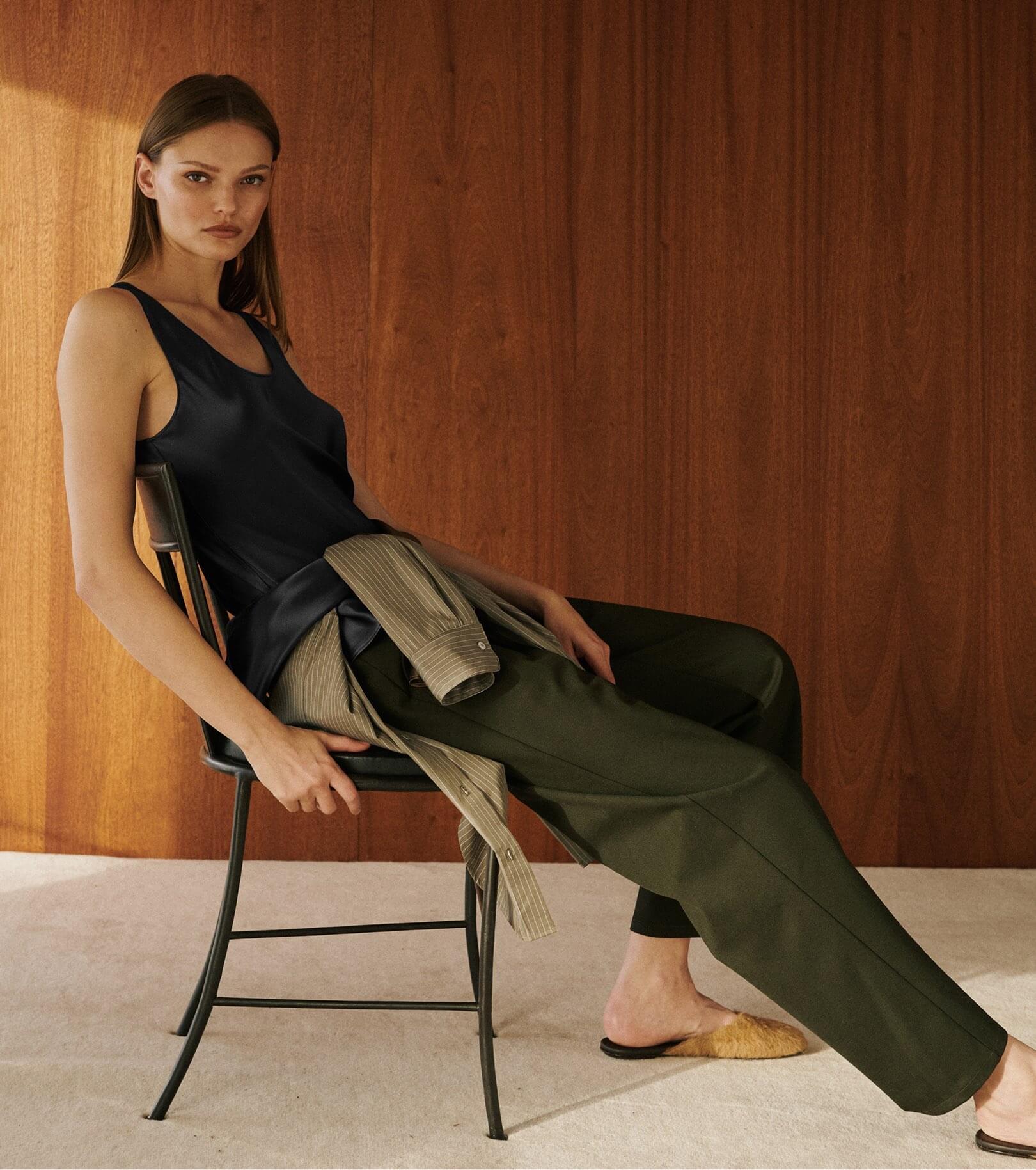 Model is sitting in chair on carpet wearing green pants and black satin tank with slides.