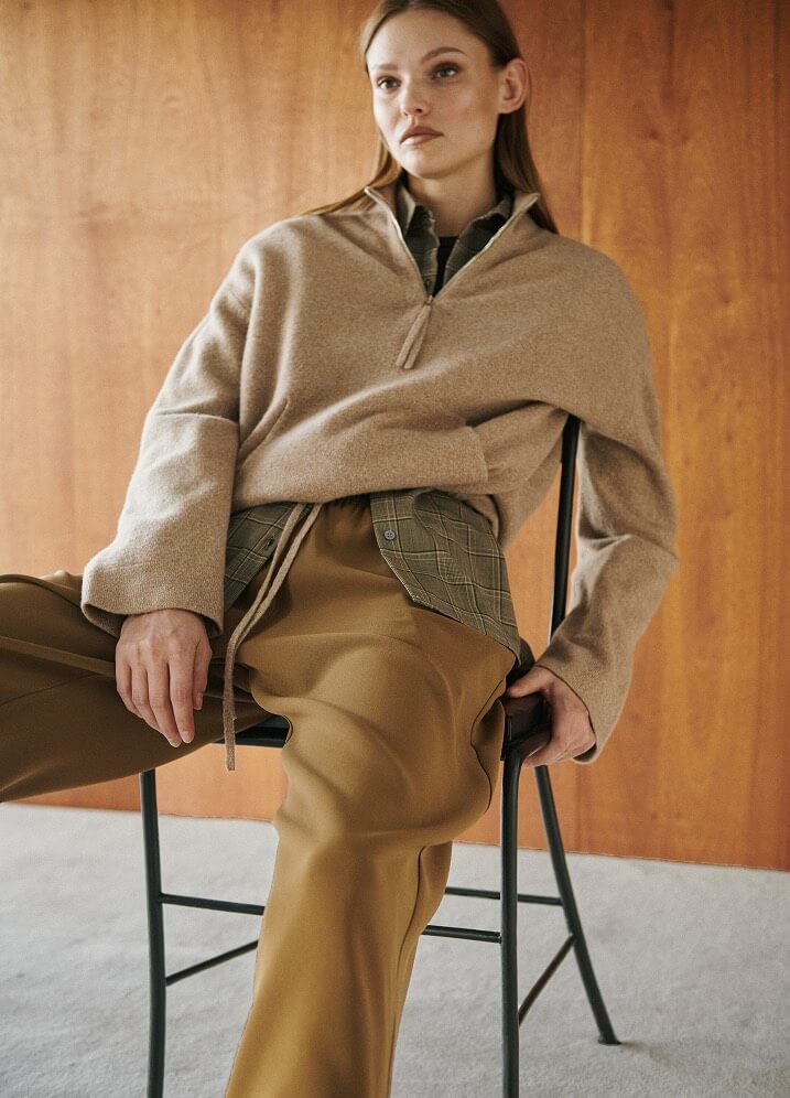 Model is wearing brown zip up sweater with flannel and brown pants, sitting in chair.