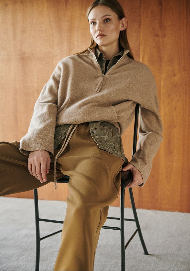 Model is wearing brown zip up sweater with flannel and brown pants, sitting in chair.