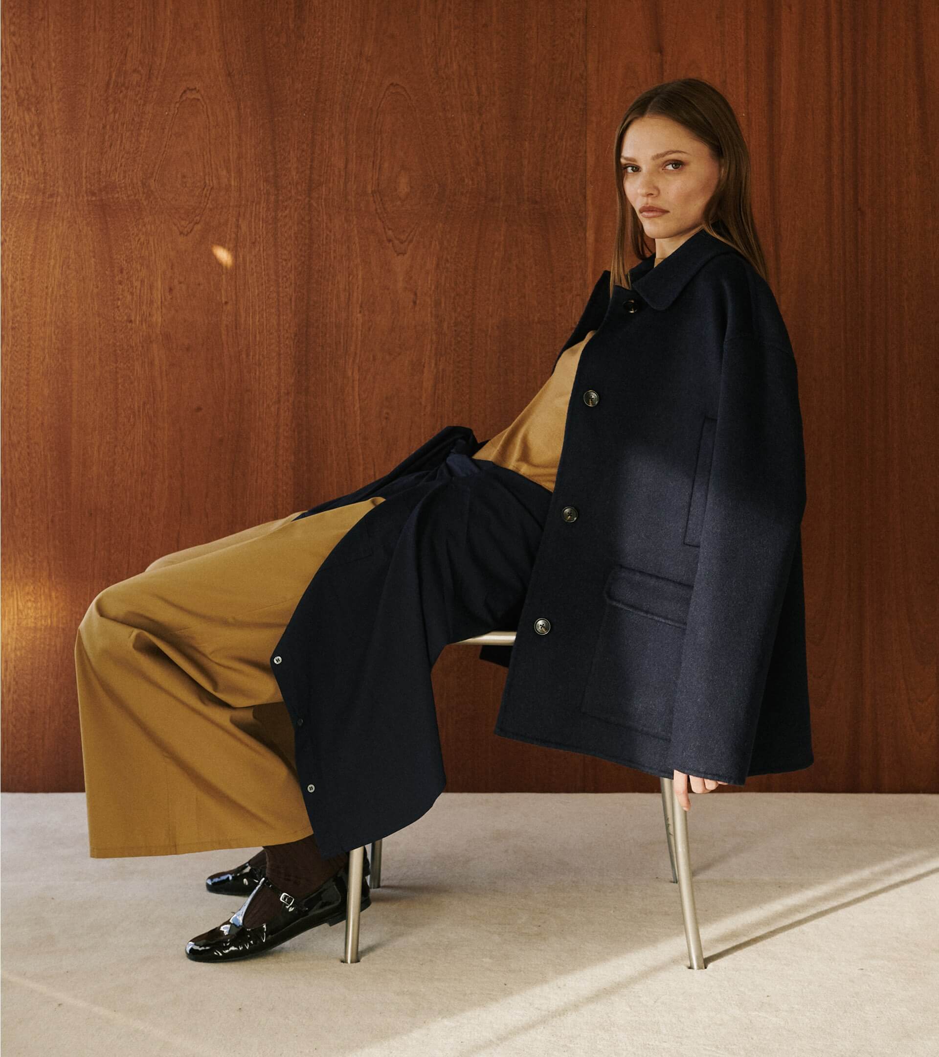 Woman sitting in chair in front of wooden wall in camel colored pants and top with coat draped over.