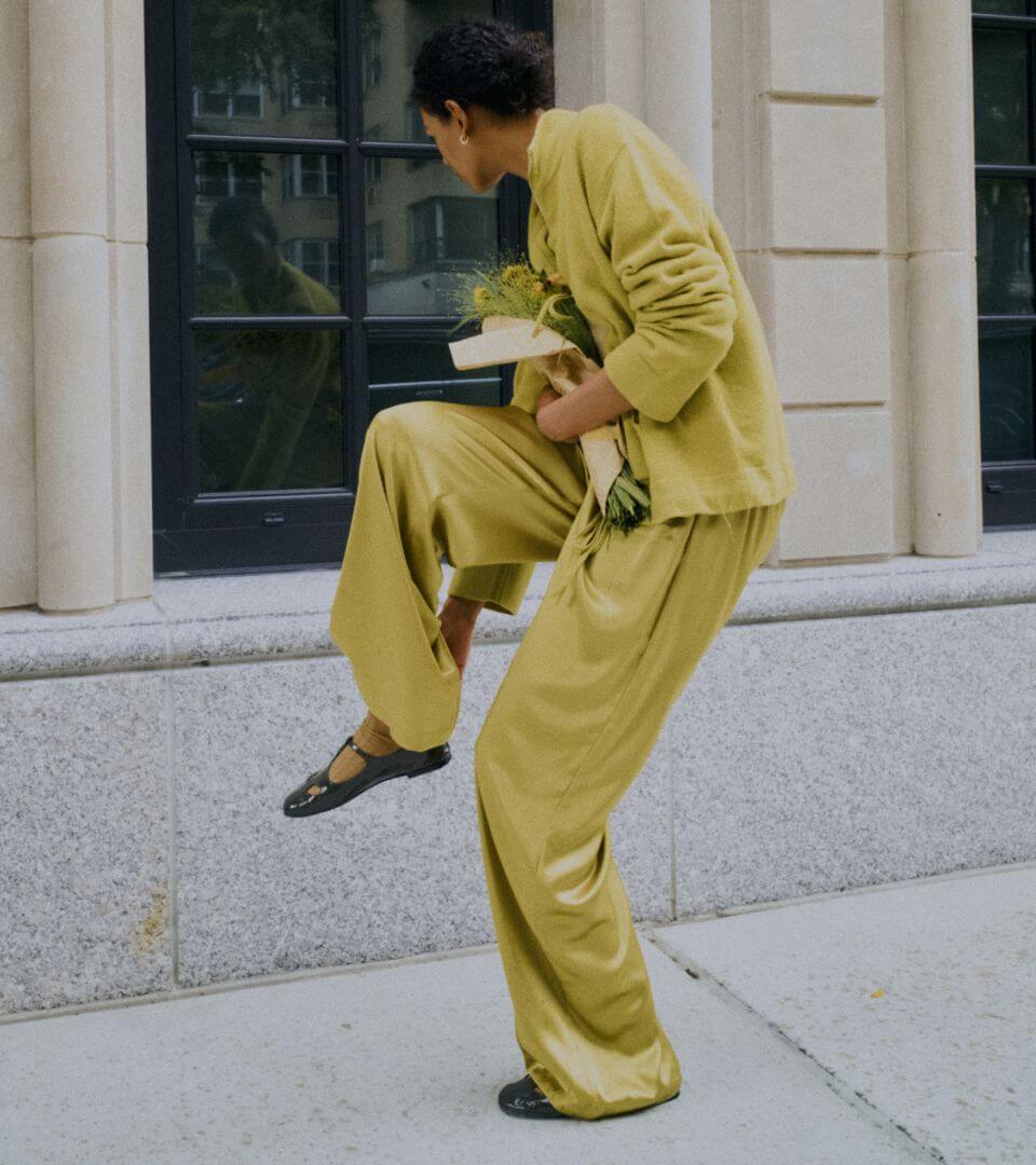 Model is standing holding shoe in green sweater and matching satin pants on sidewalk.