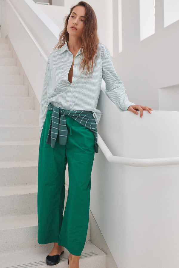 Woman standing on staircase in green Kyoto carpenter pants with Yoshi button down stripe shirt, sweater wrapped around waist.