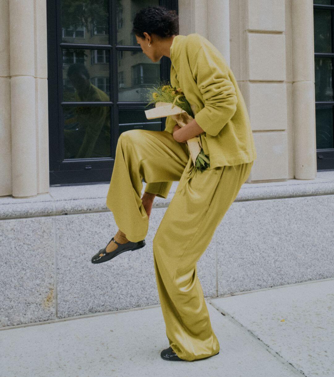 Model is standing holding shoe in green sweater and matching satin pants on sidewalk.