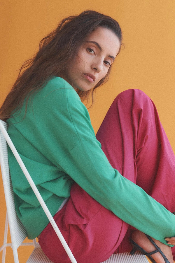 Woman sitting in white chair, in green sweater and bright pink pants, against orange wall.