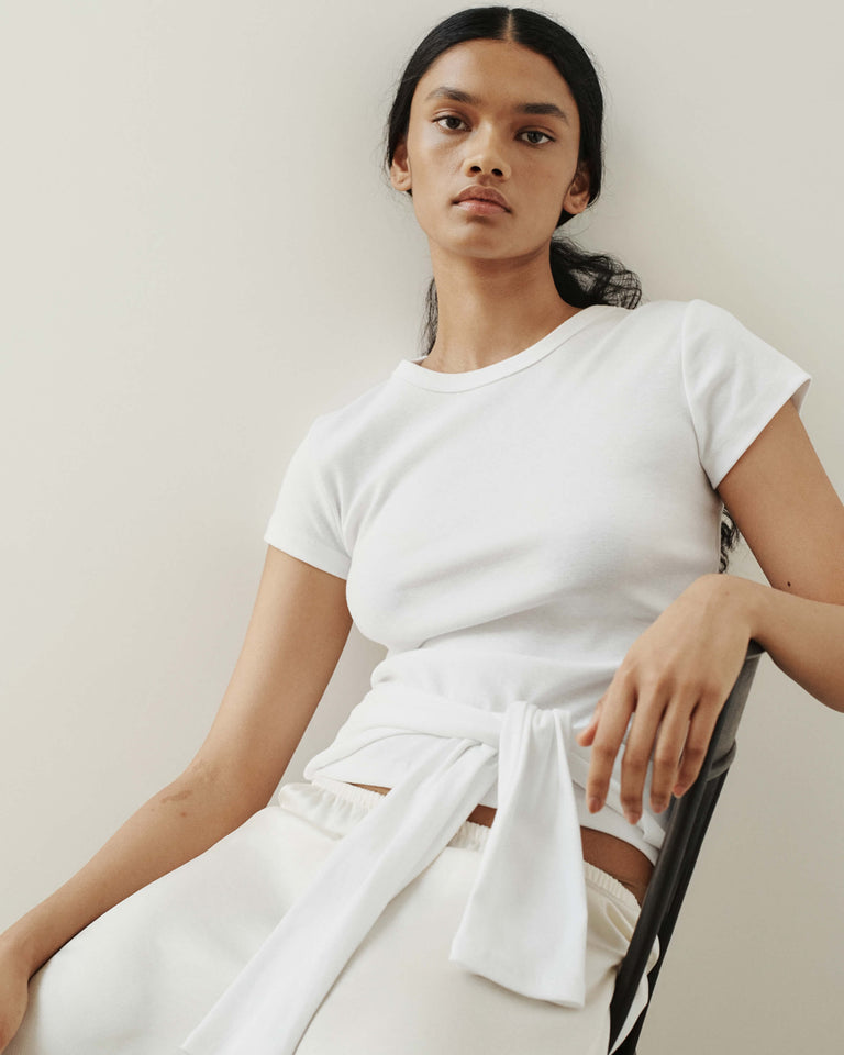 Close up of Nando white tee with white pants and sweaters wrapped around waist, woman sitting in chair against blank wall.