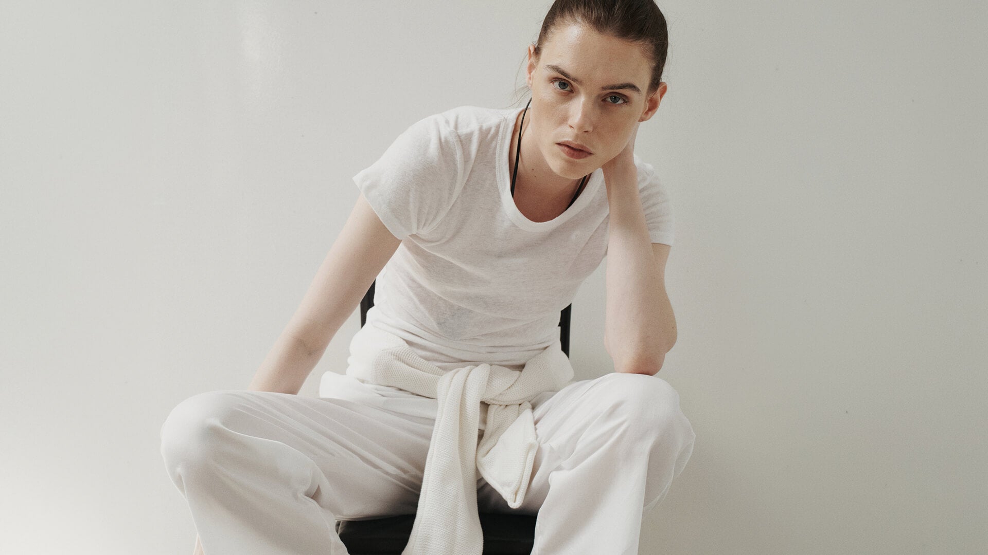 Woman sitting down wearing white tshirt and white cotton lace pants, with sweaters tied around waist, in front of blank wall.
