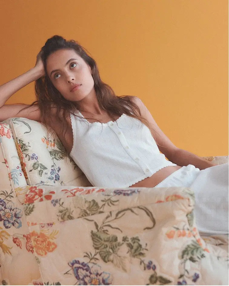 Woman sitting down in floral chair with orange background, wearing white Pointelle button down tank.