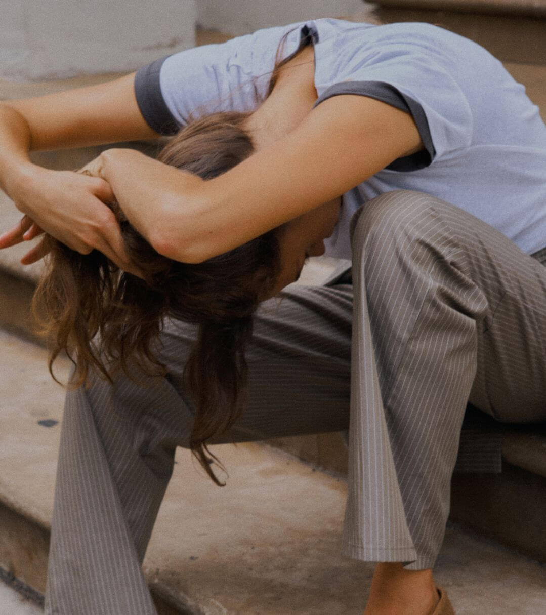 Model is leaning down with hands over head on steps, wearing t-shirt and pinstripe pants.
