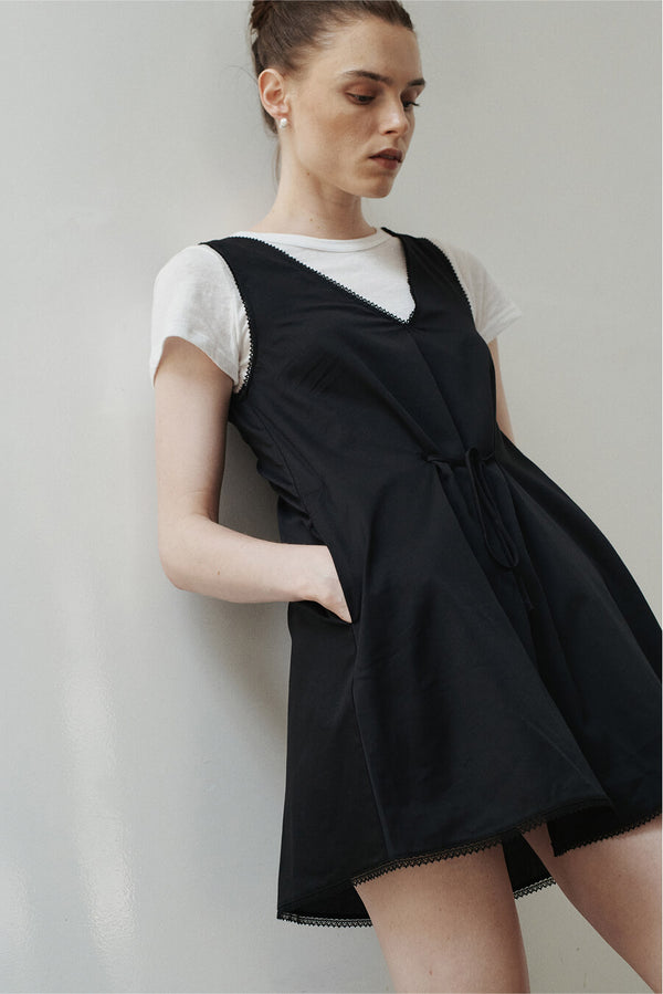 Woman wearing cotton lace black dress, layered over white tshirt learning against wall.