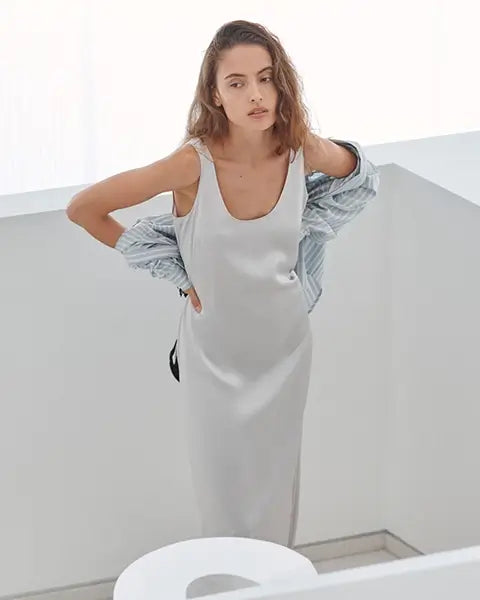 Woman standing in satin slip skirt holding heels, with brown purse and light blue tee.