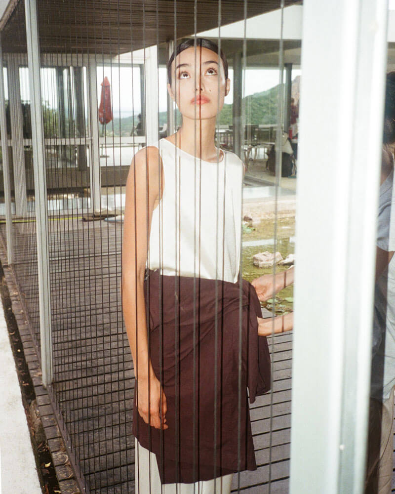 Woman in Barb satin creme set and brown sweater tied around waist, looking through window