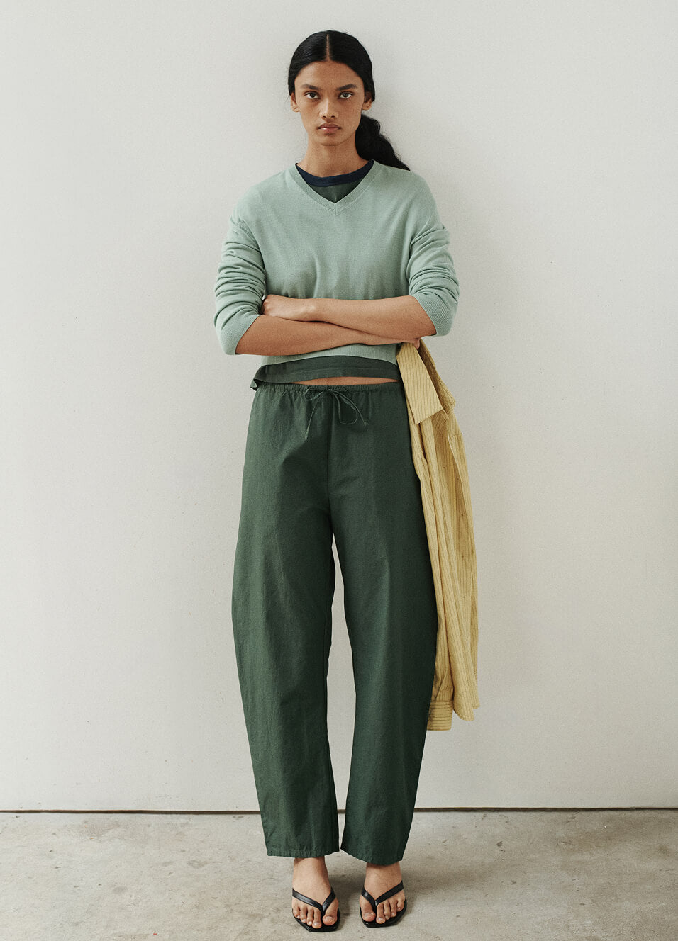 Woman standing with arms crossed in front of wall, in green carpenter pants and v neck sweater with sandals.