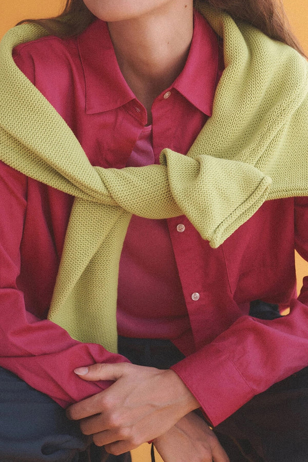 Close up of bright pink button down with green sweater draped over, arms crossed.