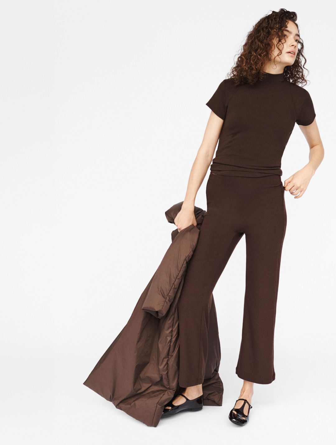 Model in chocolate lauren set, margo tee and crop flare pant with black mary jane shoes in front of white background and carrying bo anorak.