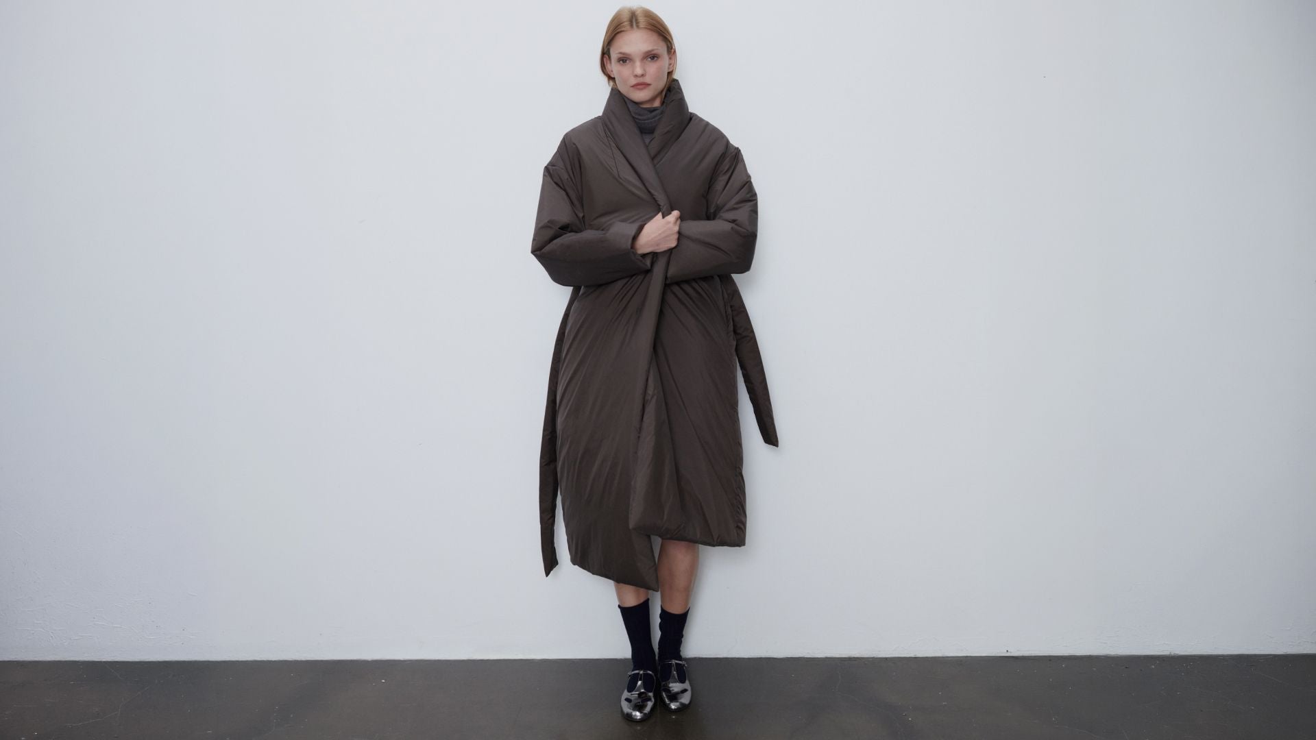 Model is wearing bo puffer shawl anorak in chocolate with socks standing in front of white wall.