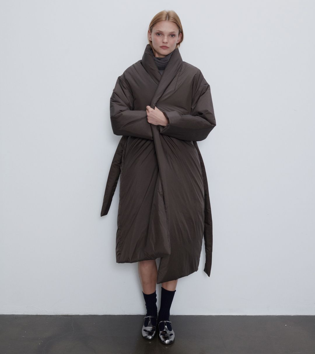 Model is wearing bo puffer shawl anorak in chocolate with socks standing in front of white wall.