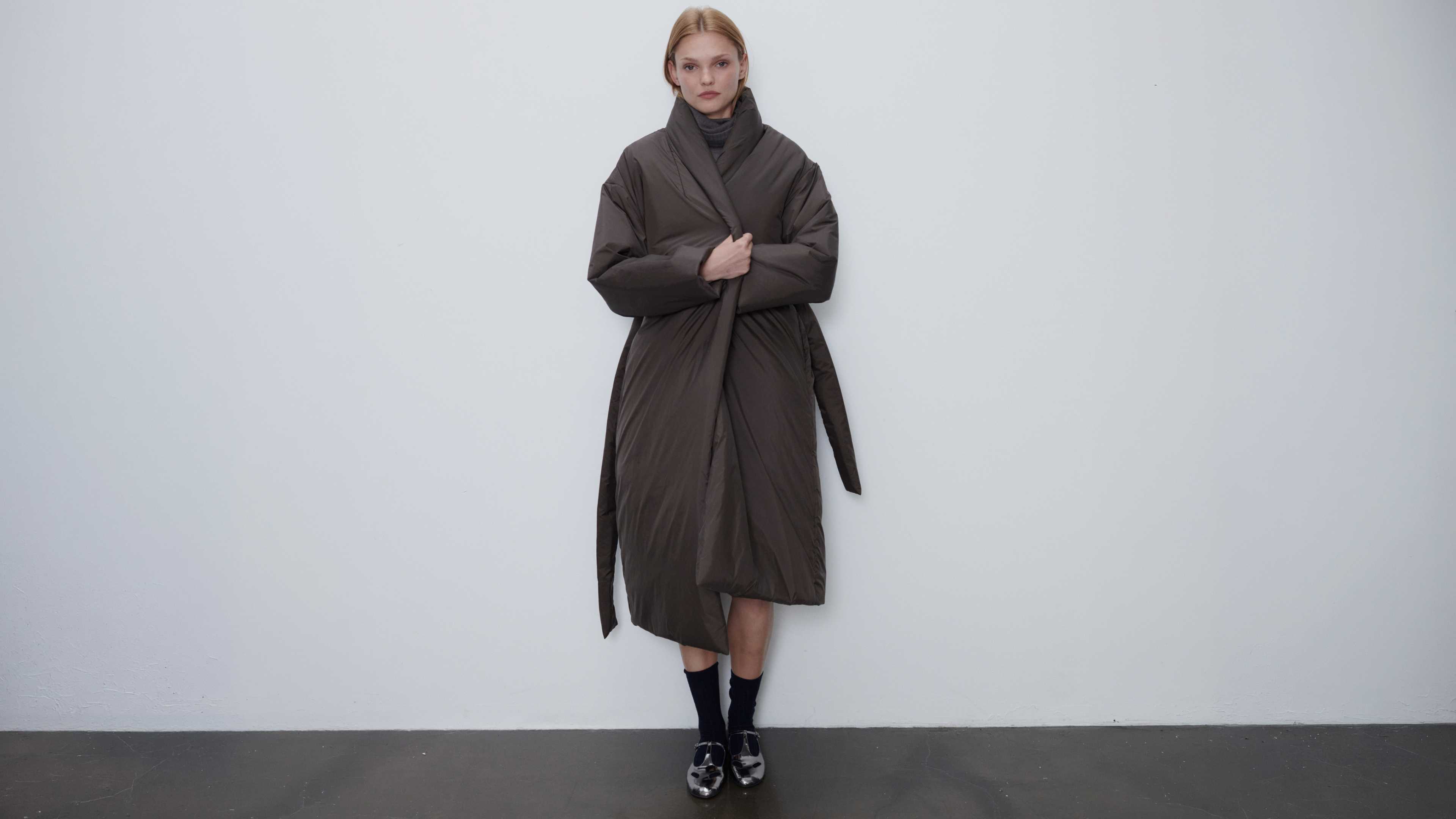 Model is wearing bo puffer shawl anorak in chocolate with socks standing in front of white wall.