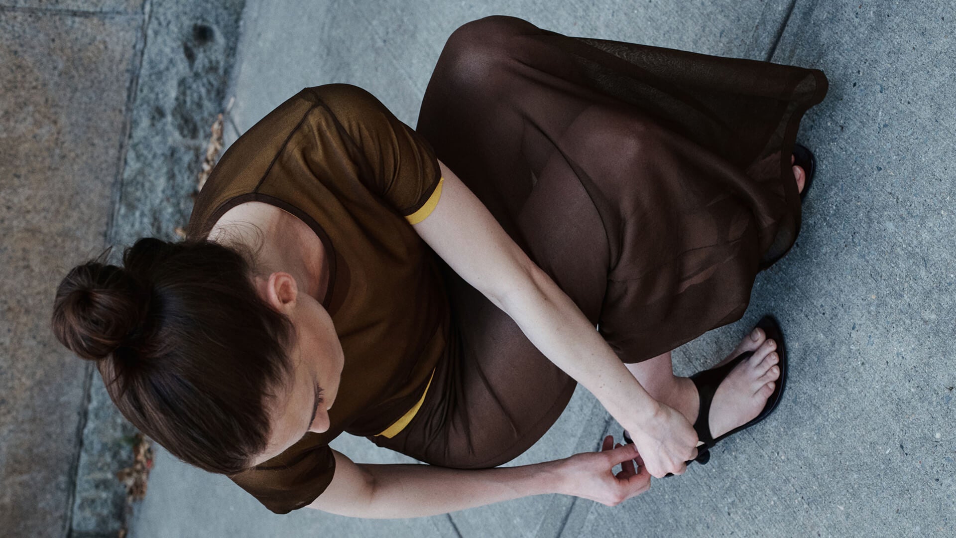 Woman sitting on sidewalk fixing shoe, in brown matching tee and skirt.