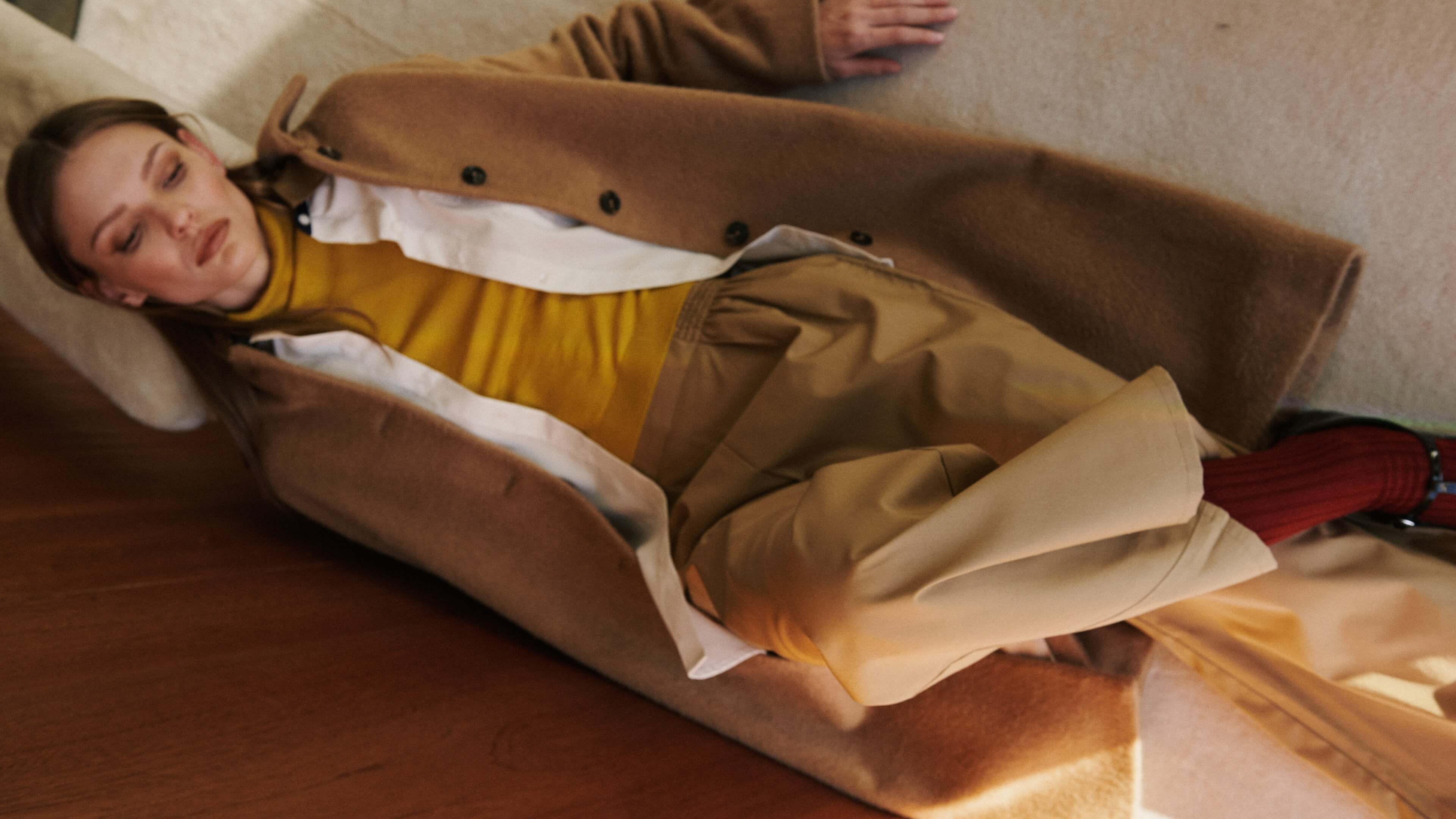 Woman laying down in coat layered over pants and white button down with yellow turtleneck.