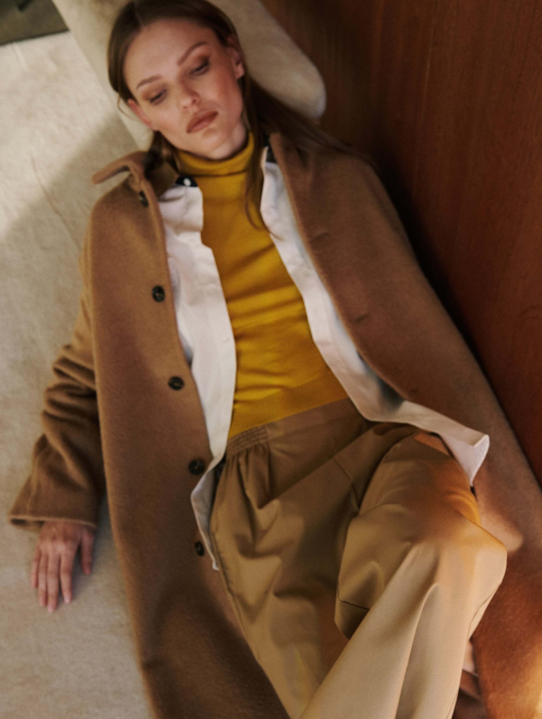 Woman laying down in coat layered over pants and white button down with yellow turtleneck.