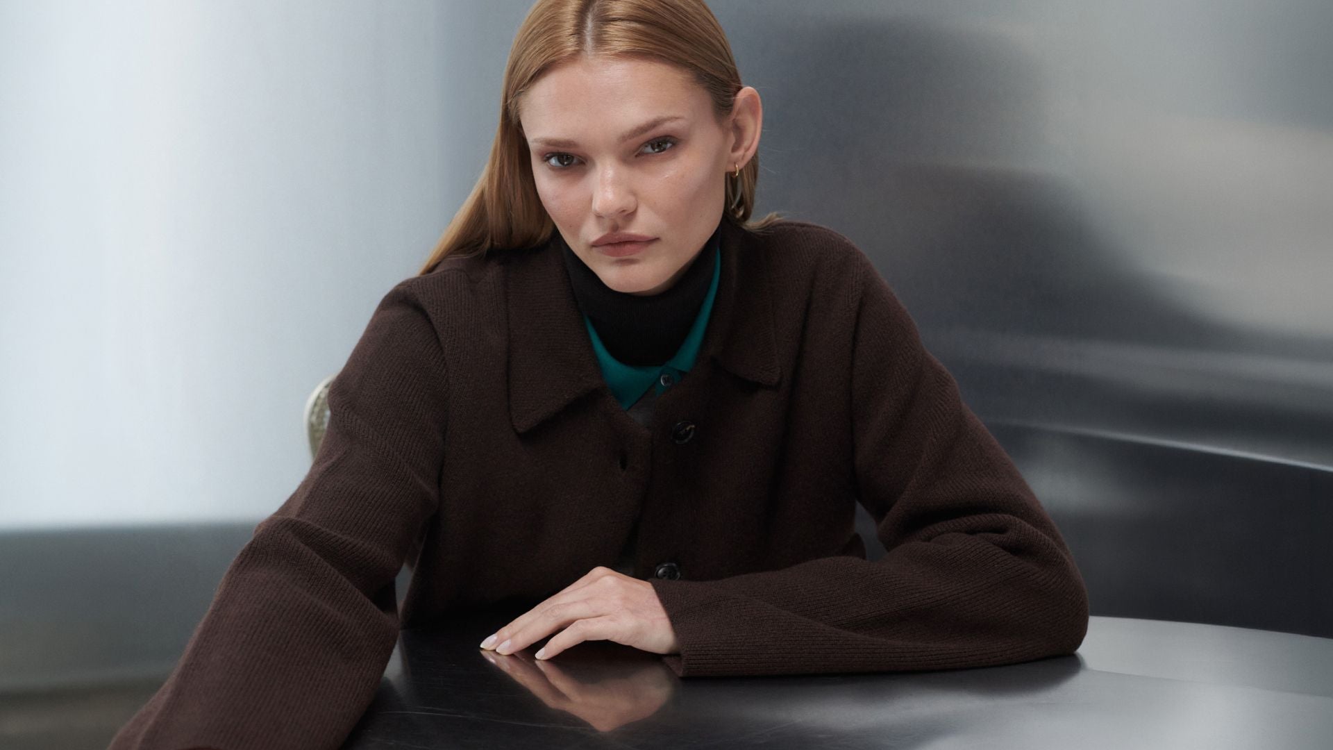 Model is leaning down on table in zoe chocolate sweater jacket layered over teal collar and turtleneck.