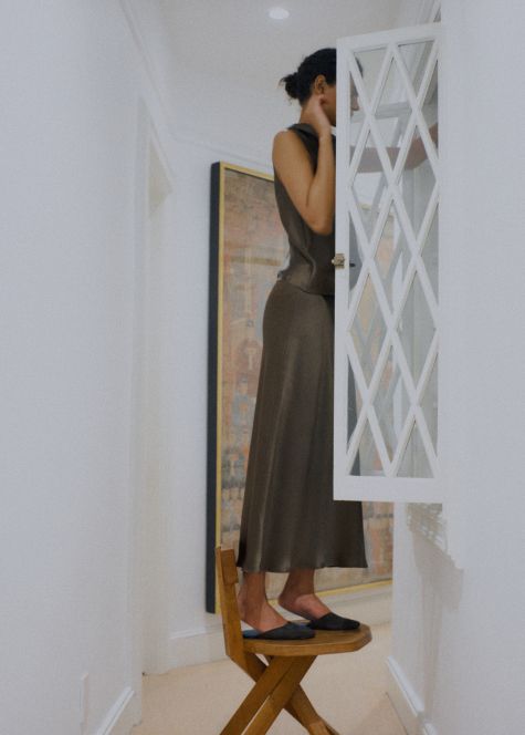 Model is standing on stool in satin set, with painting and white wall behind her, looking in cabinet.