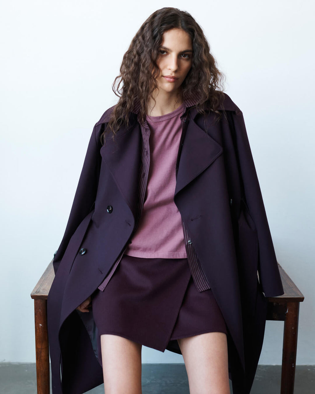 Woman in bordeaux trench layered over maud Margo Tee, sitting and leaning against a wooden table.