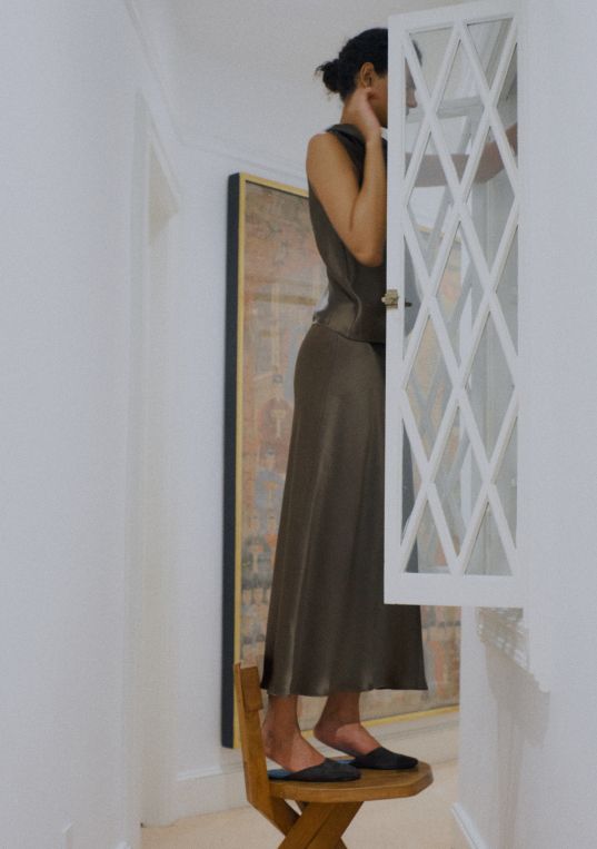 Model is standing on stool in satin set, with painting and white wall behind her, looking in cabinet.