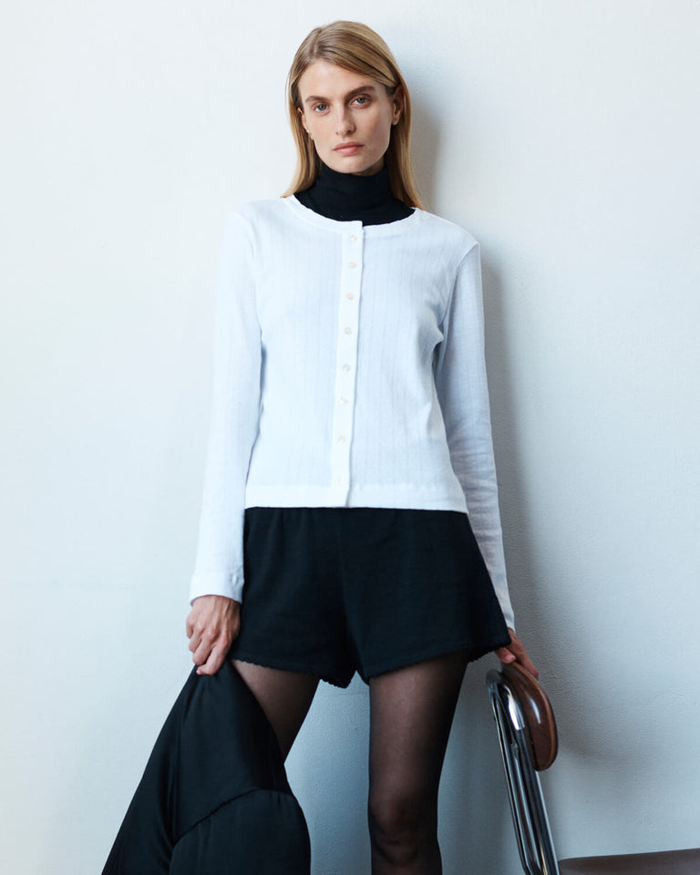 Woman in Pointelle White Cardigan and black shorts, standing against wall.