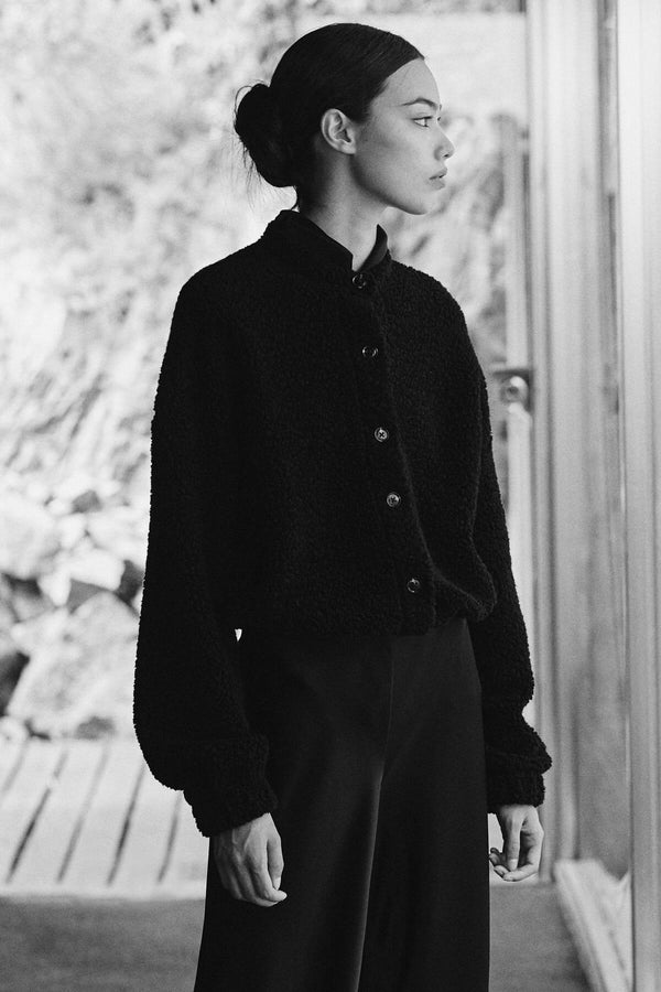 Woman standing in mica black blouson and barb satin pants, looking off into distance