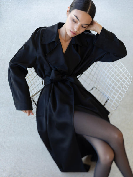 Woman in barb satin trench leaning against chair looking away